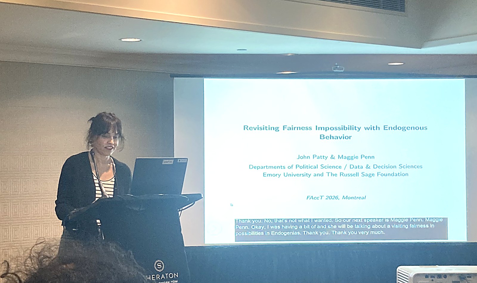 Maggie Penn at a hotel-ballroom podium at left, with the projected title slide 'Revisiting Fairness Impossibility with Endogenous Behavior' dominating the frame and a live auto-caption along the bottom.