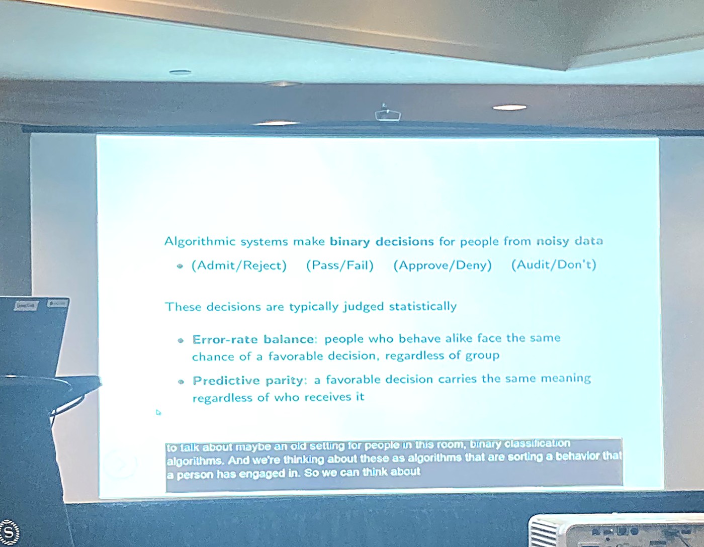 A projected conference slide showing binary decisions and the two fairness criteria, error-rate balance and predictive parity, with a live auto-caption transcribing the speaker along the bottom.